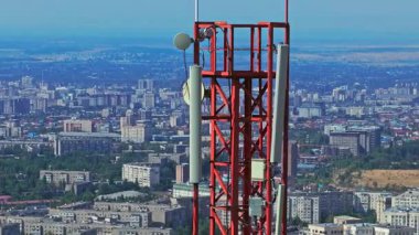 Yüksek binalar, inşaat alanları ve yerleşim alanlarının yer aldığı kalabalık bir Bişkek kentinin üzerinde panel ve çanak antenlere sahip bir telekom kulesi bulunuyor. Drone görünümü, ark hareketi.