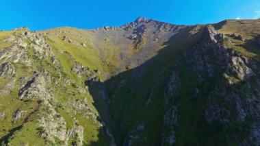 İnsansız hava aracı görüntüleri Kırgızistan 'daki dar ve kayalık bir dağ geçidini yakalıyor, sarp kayalıklar ve yemyeşil alanlarla çevrili, açık mavi bir gökyüzünün altındaki uzak bir zirveye götürüyor..