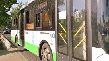 Kırgızistan, Bişkek 'te güneşli yaz gününde yeşil ağaçlarla kaplı dar Asya şehir sokağından geçen belediye otobüsü ve arabalar - 21 Haziran 2025