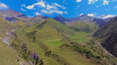 Drone captures a lush green jailoo, a high-altitude pasture in Kyrgyzstan, surrounded by rugged mountains under a partly cloudy sky. A small river flows through the valley, winding between grassy hills.