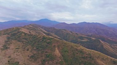 Kırgızistan 'daki Chu Vadisi' nin engebeli eteklerini görüntüleyen hava görüntüleri, kayan tepeleri, katmanlı dağları ve bulutlu gökyüzünü gösteriyor, kamera sürekli olarak doğal manzara üzerinde hareket ediyor..