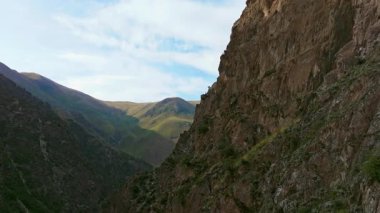 Kırgızistan 'daki dramatik bir dağ vadisini görüntüleyen hava görüntülerinde sarp kayalıklar, yeşil yamaçlar ve parçalı bulutlu bir gökyüzünün altında kayan tepeler görülüyor. Kamera yükselir, geniş araziyi gözler önüne serer..