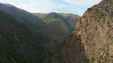 Kırgızistan 'daki dramatik bir dağ geçidini görüntüleyen hava görüntülerinde sarp kayalıklar, yeşil yamaçlar ve parçalı bulutlu bir gökyüzünün altında kayan tepeler görülüyor. Kamera ilerliyor..