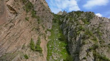 Kırgızistan 'daki engebeli bir dağ oluğu boyunca, dik kayalıkların arasından geçen doğal kayalık bir yolu ve açık mavi gökyüzünün altındaki seyrek yeşilliği gösteren bir uçuş görüntüsü..