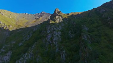Drone görüntüleri Kırgızistan 'daki dramatik bir dağ vadisini, kayalık kayalıkları, yeşil yamaçları ve açık mavi gökyüzünün altında uzak bir zirveyi yakalar. Kamera sağa sola hareket ediyor, parallaks boyunca derinliği gösteriyor.