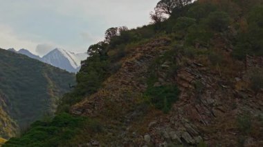 Drone görüntüleri Kırgızistan 'da güneş doğarken ya da batarken çarpıcı bir dağ vadisini yakalıyor. Çekim, yemyeşil yamaçlar, kayalık uçurumlar ve parçalı bulutlu gökyüzünün altındaki uzak karlı tepeleri ve güneş ışığının ılık bir parıltıyı yansıtıyor..