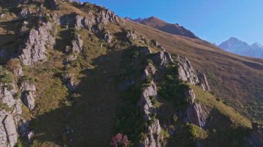 Kırgızistan 'daki kayalık kayalıklarla dolu bir dağ yamacının hava görüntüsü, kamera ufuk çizgisindeki karla kaplı tepeleri gösteriyor. Güneşli bir akşam, açık bir gökyüzü.