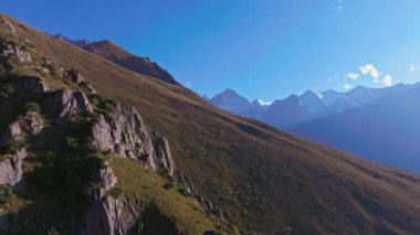 Bir dron, kayalık kayalıklar, çimenli yamaçlar ve açık mavi gökyüzünün altındaki uzak kar örtülü zirvelerin yer aldığı Kırgızistan engebeli dağ manzarasının nefes kesici hava görüntüsünü yakalar..