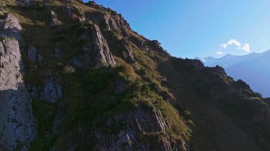 Kırgızistan 'daki kayalık kayalıklarla dolu bir dağ yamacının hava görüntüsü, kamera açık mavi gökyüzü ile güneşli bir akşamda ufukta karla kaplı tepeleri gösteriyor..