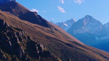 Kırgızistan 'daki dağ manzarasının hava görüntüleri. Dik, kayalık bir yamaç ön plana hükmediyor, dağların kıyısındaki bir vadiye iniyor. Uzakta karla kaplı zirveler dağınık bulutlu açık mavi bir gökyüzüne karşı.