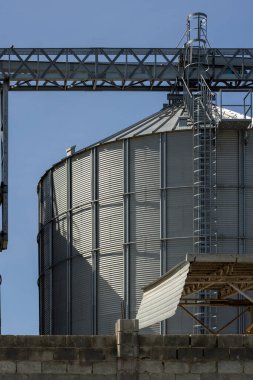 Grain silo stands tall in an industrial area under a bright sunny sky. Metal structure features stairs and a loading platform, evoking a stark and quiet atmosphere.