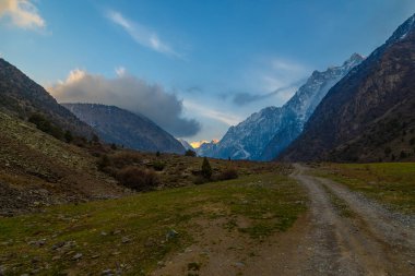 Vast mountain range dominates the horizon, while a dirt road winds through a peaceful valley. Natural environment during springtime evening evokes feelings of tranquility and awe in Kyrgyzstan.