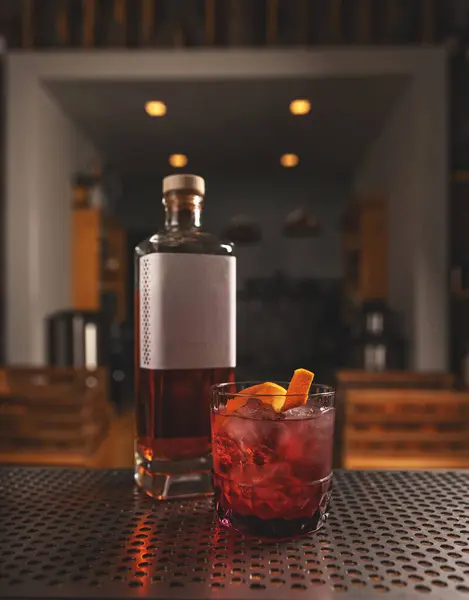 Elegantes Glas Roter Cocktail Mit Eis Und Orangenschale Neben Whiskeyflasche lizenzfreie Stockfotos