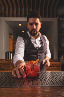 Yelek giymiş uzman bir barmen. Yeni yapılmış negroni kokteyli ve portakal kabuğu garnitürü sunuyor.