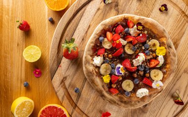 Üzerinde taze meyve ve böğürtlen bulunan tatlı bir pizzanın kırsal ahşap bir masada manzarası.