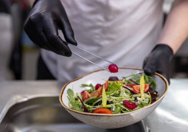 Şef, restoranın mutfağındaki renkli bir salataya soğan turşusu eklemek için maşa kullanıyor. Sağlıklı ve lezzetli bir yemek hazırlıyor.
