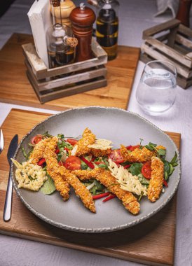 Çeri domatesli ve parmesan peynirli taze yeşil salatanın üzerinde nefis ekmeklenmiş tavuk şeritleri servis edilir.