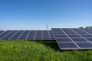 Almanya 'da arkada elektrik hatları ve rüzgar türbinleri olan güneş santrali görülüyor.