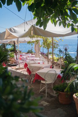 Deniz kıyısındaki yazlık kafe. Büyük yeşil çalıların sıcak güneşinden gelen tentenin altında beyaz masa örtüleri. Sıcak bir yaz gününde dinlenin. Amalfi, İtalya, deniz manzarası