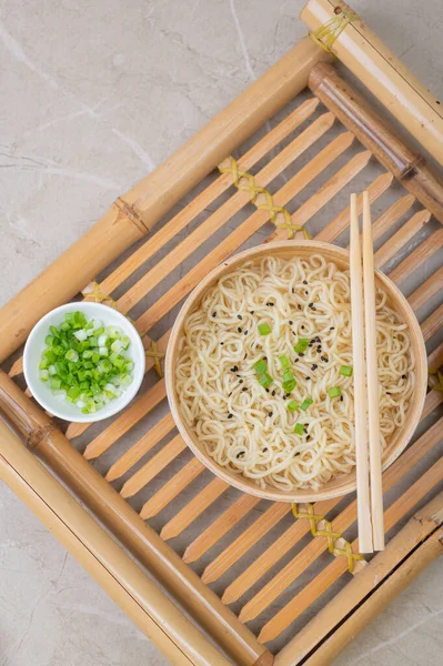 White bamboo plate (bowl) with egg noodles and plates with ingredients for it on a bamboo serving tray against a light background. Asian traditional fast food