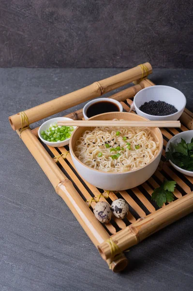 White bamboo plate (bowl) with egg noodles and plates with ingredients for it on a bamboo serving tray on a dark marble (slate) background. Asian traditional fast food