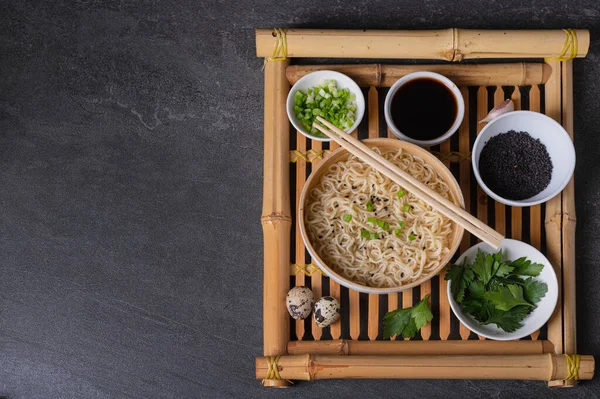 White bamboo plate (bowl) with egg noodles and plates with ingredients for it on a bamboo serving tray on a dark marble (slate) background. Asian traditional fast food