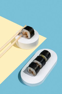 Hosomaki (suşi, rulo) beyaz bir alçı üzerinde çok renkli düz bir zemin üzerinde yılan balığı ile (mavi, sarı). Basit bir özet kompozisyon