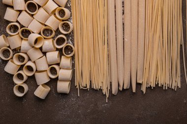 Kahverengi arka planda el yapımı makarna bu fotoğrafın konusu. Erişteler çeşitli şekil ve boyutlarda ve taze ve pişmemiş görünüyor. Buğdaydan ev yapımı makarna.