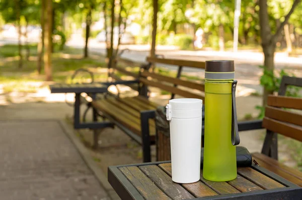 ウォーターボトルと魔法瓶カップは絵のように美しい公園の設定でテーブルの上に置かれ 容器を再利用し 持続可能性を促進することの重要性を強調しています 環境に優しい習慣 — ストック写真