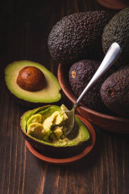 Kil tabakta avokado, karanlık ve kasvetli bir ahşap zemine karşı. Bu modaya uygun kompozisyon görsel olarak çekici bir karşıtlık yaratır. Yarım kaşıkla avokadoyu kazıdım. Sağlıklı beslenme, Karanlık Huysuz tarzı.