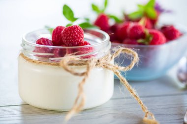 Cam bir kavanozda taze ahududu bulunan ev yapımı yoğurt, açık ahşap bir masada..