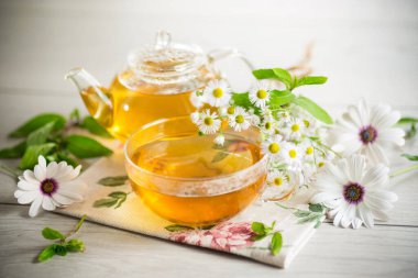 Papatya çiçeği çayı cam fincanda ve çaydanlıkta, açık ahşap bir masada..