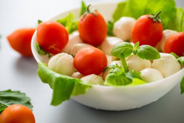Beyaz kâse içinde mini mozarella otlar ve gri arka planda vişneli domatesler..