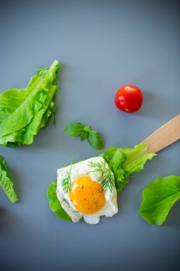 İştah açıcı sahanda yumurta, ahşap spatulada yeşiller, gri arka planda..