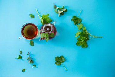 Frenk üzümü yapraklı yeşil çay ve cam çaydanlıkta nane ve mavi arka planda fincan. Üst görünüm .