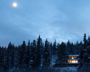 Cozy country living home warmly illuminated isolated in boreal forest taiga moon-lit frozen winter landscape of remote Yukon Territory, YT, Canada clipart