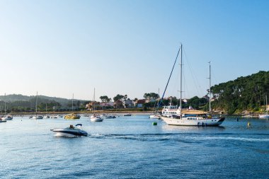 PONTEVEDRA, İspanya - 24 Haziran 2023: Güneşli bir yaz gününde Portonovo yat kulübüne (Pontevedra, İspanya) demirlemiş küçük tekneler, limana giren küçük bir yatla.