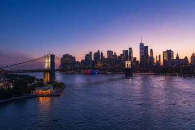 Brooklyn Köprüsü ve New York Şehri East River 'ın karşısındaki ufuk çizgisi yoğun ve yakıcı bir günbatımıyla aydınlandı. Gökyüzünün dramatik renkleri suyu yansıtıyor, köprünün mimari ihtişamını ve çarpıcı ikonik şehir manzarasını vurguluyor.