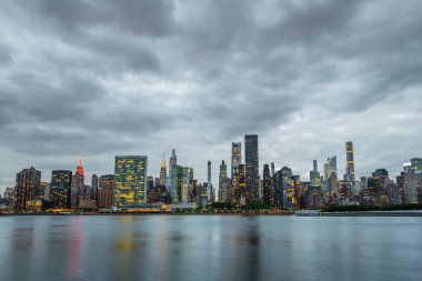 New York 'taki Long Island City' deki Gantry Plaza Eyalet Parkı 'ndan Manhattan' ın ufuk çizgisi manzarası. Uzun pozlama, şehrin ikonik gökdelenlerinin kasvetli, bulutlu gökyüzüne karşı dramatik bir kontrastını yakalıyor ve dinamik ve büyüleyici bir şehir yaratıyor.