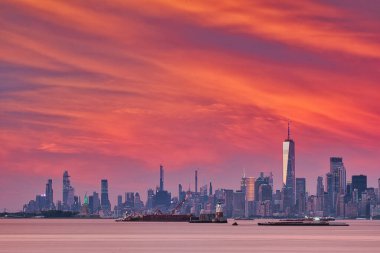 Gün batımında Manhattan ufuk çizgisinin göz kamaştırıcı manzarası, canlı turuncu ve pembe renkler gökyüzünü aydınlatıyor. İkonik bir Dünya Ticaret Merkezi diğer gökdelenler arasında dimdik dururken Özgürlük Anıtı uzaktan görülebiliyor..