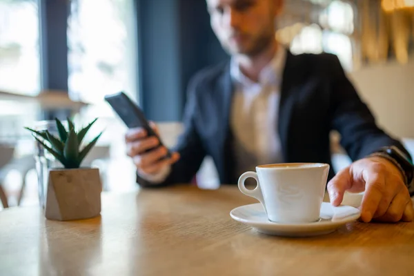 大きな白いカップから朝のコーヒーを飲み 携帯電話でニュースを読むビジネスマン コーヒーカップに焦点を当て ぼやけている — ストック写真