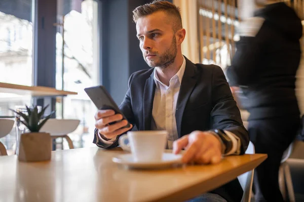 スマートフォンでニュースを読み 大きな白いカップから朝のコーヒーを飲む素晴らしい若いビジネスマン コーヒーカップがぼやけている 男に焦点を当てる — ストック写真