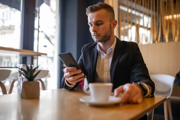 スマートフォンでニュースを読み 大きな白いカップから朝のコーヒーを飲む素晴らしい若いビジネスマン コーヒーカップがぼやけている 男に焦点を当てる — ストック写真