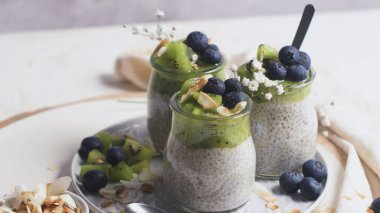 Kivili Chia pudingi, yabanmersini ve hindistan cevizi dilimleri, beyaz bir masada cam kavanozlarda üç porsiyon..