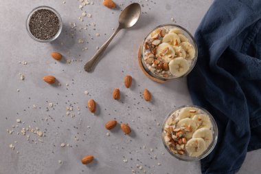 Muz dilimleri ve bademlerle kaplı iki kase yulaf. Bir kaşık, kesme tahtası, bıçak ve yanında mavi bir peçete..