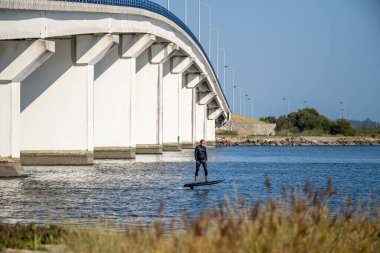 A lone hydrofoil surfer glides beneath a massive white bridge, blending urban architecture with watersport excitement on a calm river clipart