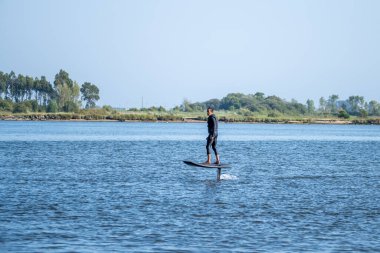 Yalnız bir sörfçü, berrak bir günde, ağaçlardan ve kıyılardan oluşan sakin bir manzarayla çevrili bir su folyosunun üzerinde süzülür.