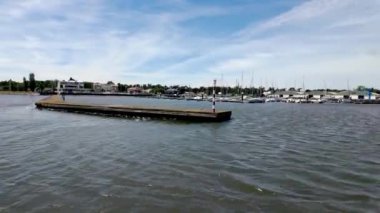 Deniz manzaralı sakin sular, nehre uzanan bir rıhtım ve güneşli bir günde parçalı bulutlu bir gökyüzünün altında arka planda yelkenliler.