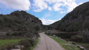 Buracas do Casmilo, Condeixa, Portekiz yakınlarındaki kayalık yamaçlı dar bir vadiden geçen toprak yol. Zeytin ağaçları ve dağınık bulutlu mavi gökyüzünün altında dağılmış taş duvarlar..