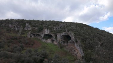 Buracas do Casmilo at Condeixa - Portugal. Ancient limestone caves nestled in a rugged hillside, featuring multiple natural cavern openings amid green vegetation and moss-covered rock formations on a cloudy day. clipart